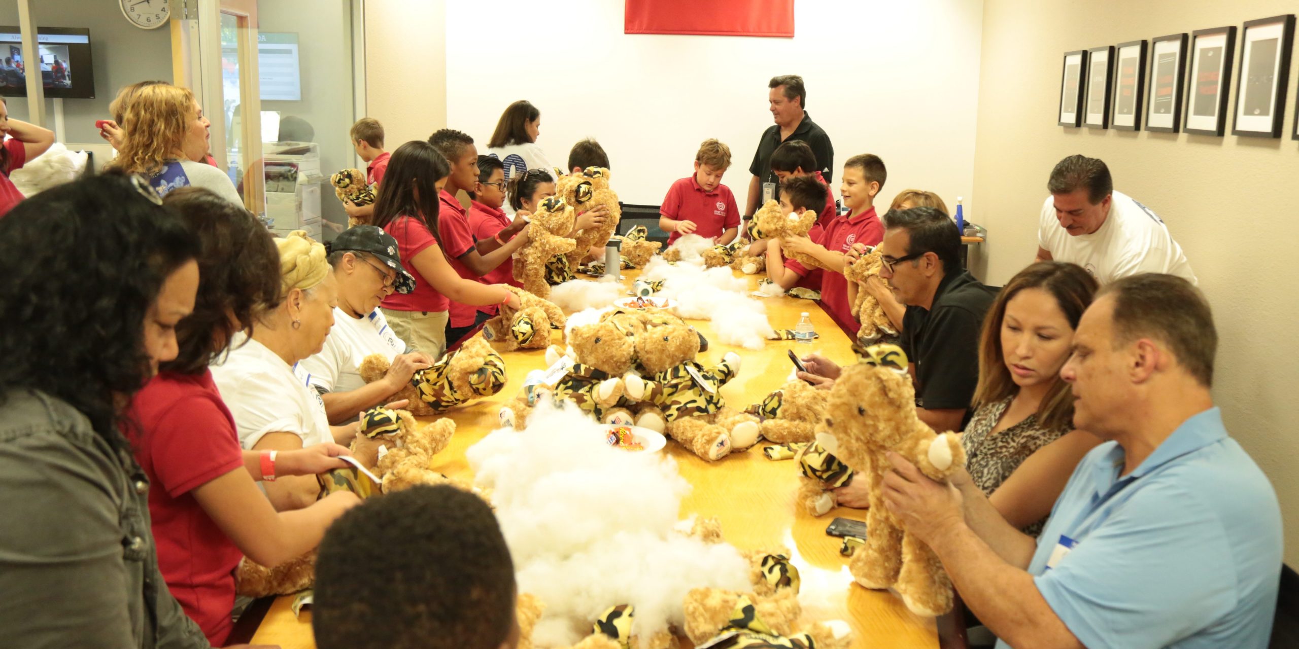 A diverse group of people, including children and adults, sit around a long table at a Bear Building Event, assembling stuffed animals with plush toys and stuffing spread out. The atmosphere is busy and collaborative.