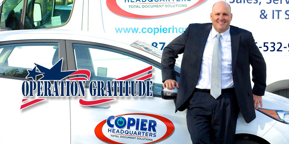 A man in a suit smiles while leaning on a car with a Copier Headquarters logo, representing the Los Angeles firm. Behind him is a branded van. The text Operation Gratitude with a star and flag graphic is overlaid on the image.