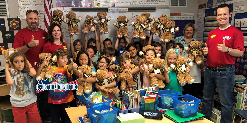 A group of smiling children and adults in a colorful classroom hold up teddy bears. Artwork decorates the walls, books fill the desks, and an Operation Gratitude banner is proudly displayed in the foreground.