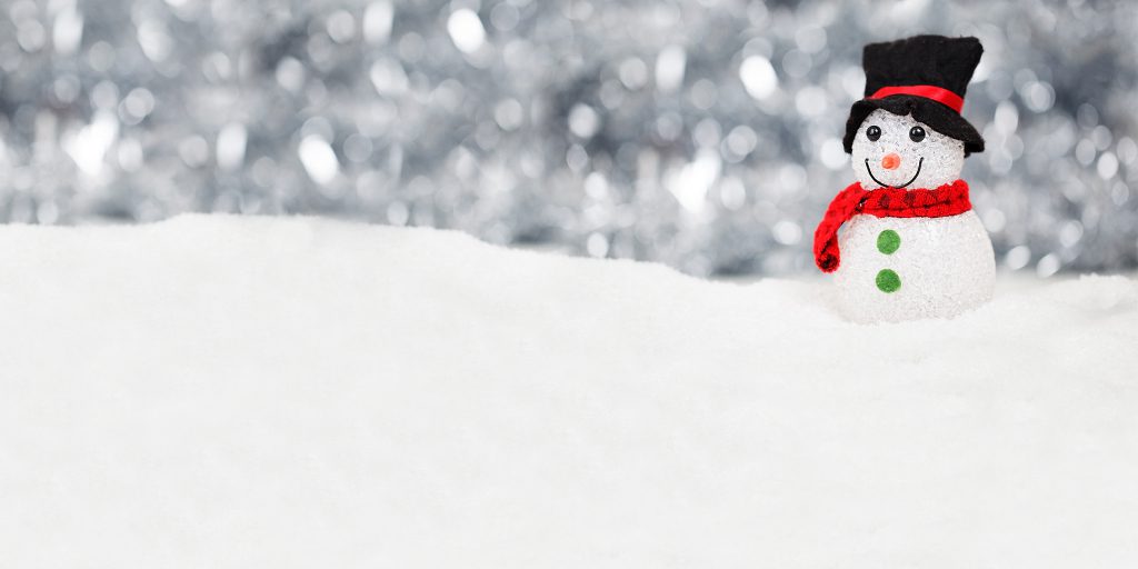 A small snowman with a black top hat, red scarf, green buttons, and a smiling face stands on a snowy surface with a blurred, sparkling silver background, capturing the festive spirit of Holiday Drive 2019.