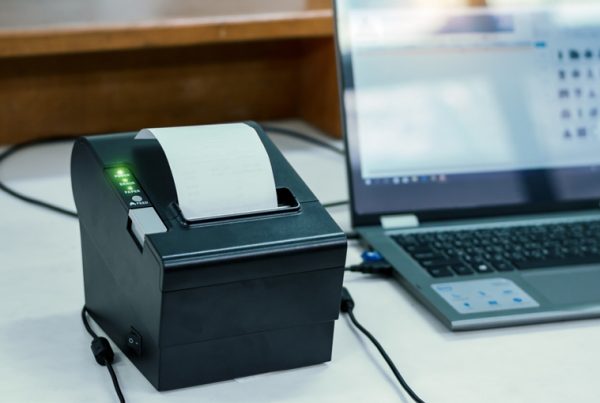 A black thermal receipt printer with a printed receipt sits on a desk next to an open laptop. Both devices are plugged in and connected, showcasing a great choice for anyone consulting a printer buying guide.