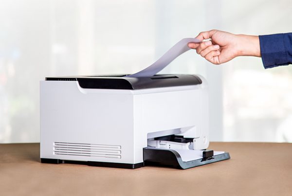 A person’s hand is placing a sheet of paper into a white office printer on a brown surface, with a blurred light background.