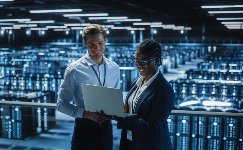Two professionals stand in a large, modern server room, smiling and looking at a laptop as they conduct a cybersecurity assessment. Rows of illuminated server racks fill the background, creating a high-tech atmosphere.