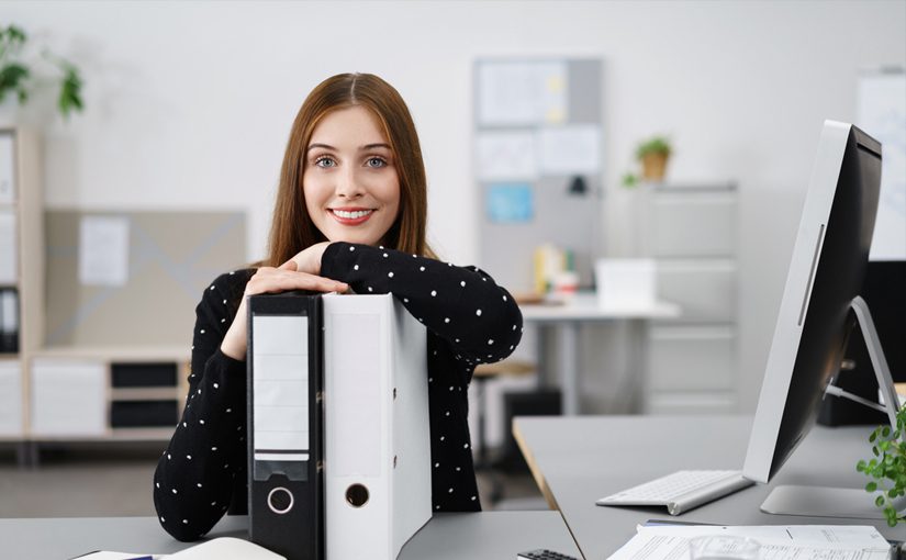 A woman with long brown hair, wearing a black polka dot sweater, sits at a desk in an office, smiling and resting her arms on two large binders—showcasing her expert office organization tips for 2023—with a computer monitor beside her.