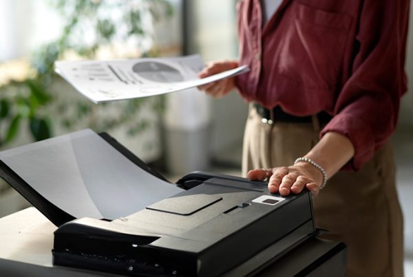 A person in a red shirt stands next to a large printer, holding printed papers and pressing a button—showcasing the ease and efficiency of Managed Print Services, with plants and sunlight visible in the background.