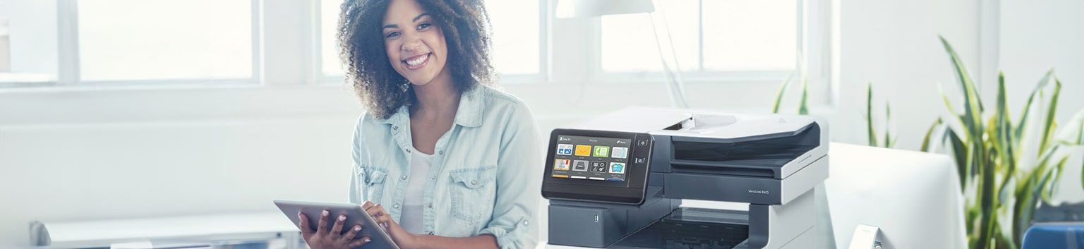 A smiling woman with curly hair holds a tablet while sitting next to a modern business printer in a bright, well-lit workspace with plants and large windows in the background.