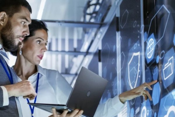 Two professionals stand in a server room, analyzing data icons on a large transparent digital screen. One holds a laptop while the other points at the display, illustrating collaboration in Managed IT Services.