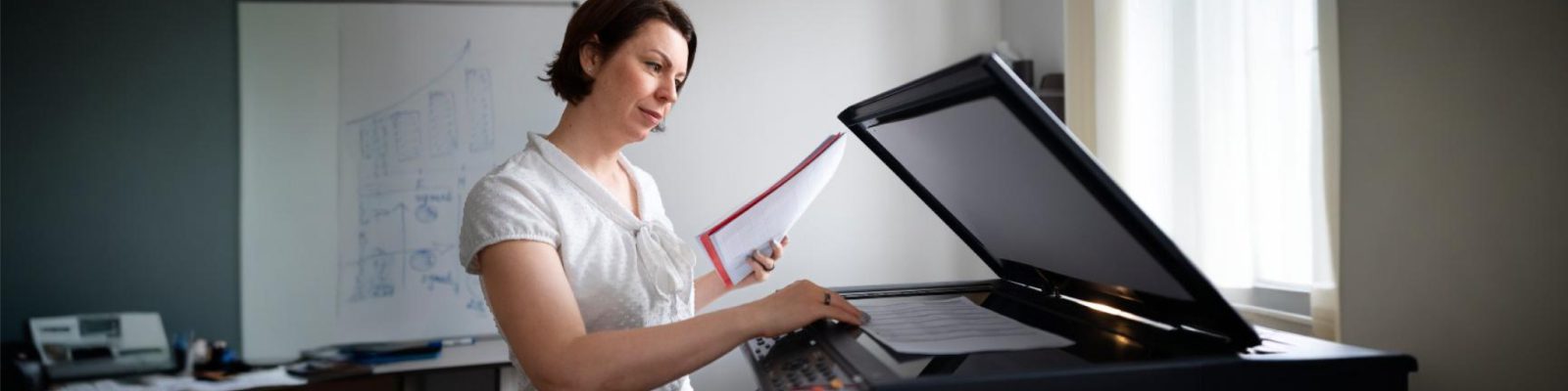 A woman stands at a photocopier, holding papers and pressing buttons, in a bright office with a whiteboard in the background—perfect for business printing needs or anyone searching for reliable Fresno printers.