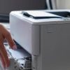 A person stands at a desk, loading paper into a white printer—ideal for small businesses—with another black printer and a computer monitor nearby, showcasing options among the top 10 commercial printers.
