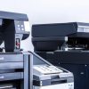 A row of modern 2025 office printers and copiers lines a white wall, each with open trays and control panels visible in the brightly lit room.