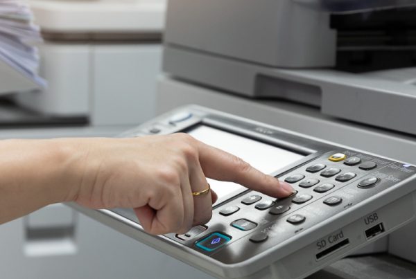 A person presses buttons on a copier machine’s control panel while holding a stack of paper in the other hand, preparing to make copies—essential for real estate agents managing important documents.