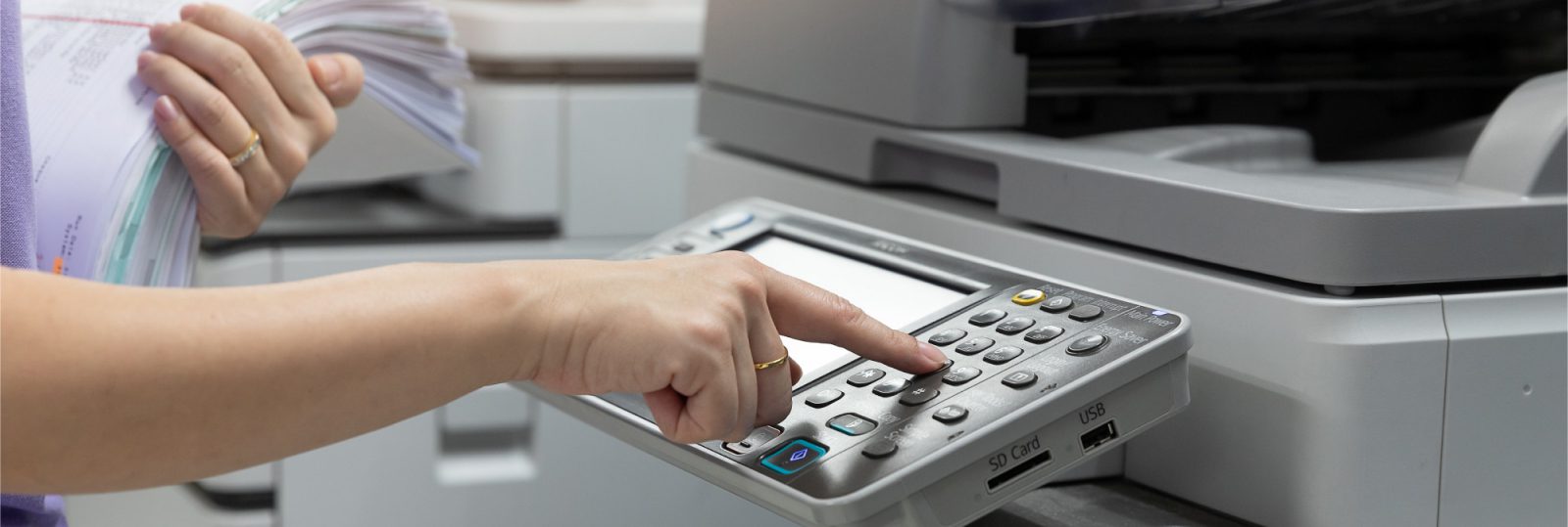 A person presses buttons on a copier machine’s control panel while holding a stack of paper in the other hand, preparing to make copies—essential for real estate agents managing important documents.