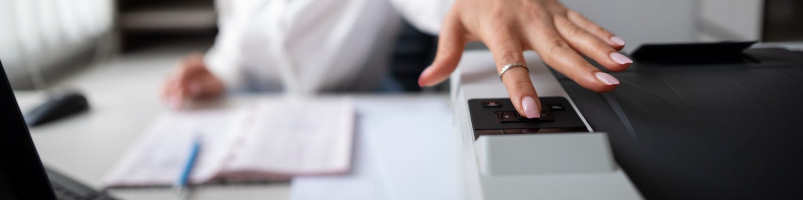 A person with manicured nails and a ring presses a button on a photocopier, often used by construction businesses, with paperwork and a pen visible on the desk in the background—highlighting the importance of commercial printers.