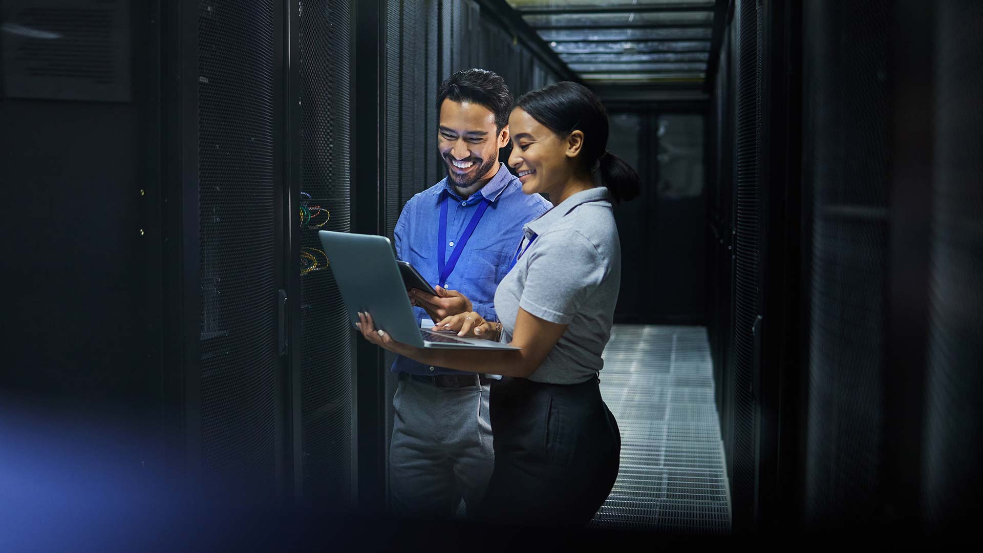 Two IT professionals stand in a server room, smiling while discussing a network assessment on a laptop. Server racks are visible on both sides, and the environment appears well-lit and modern.